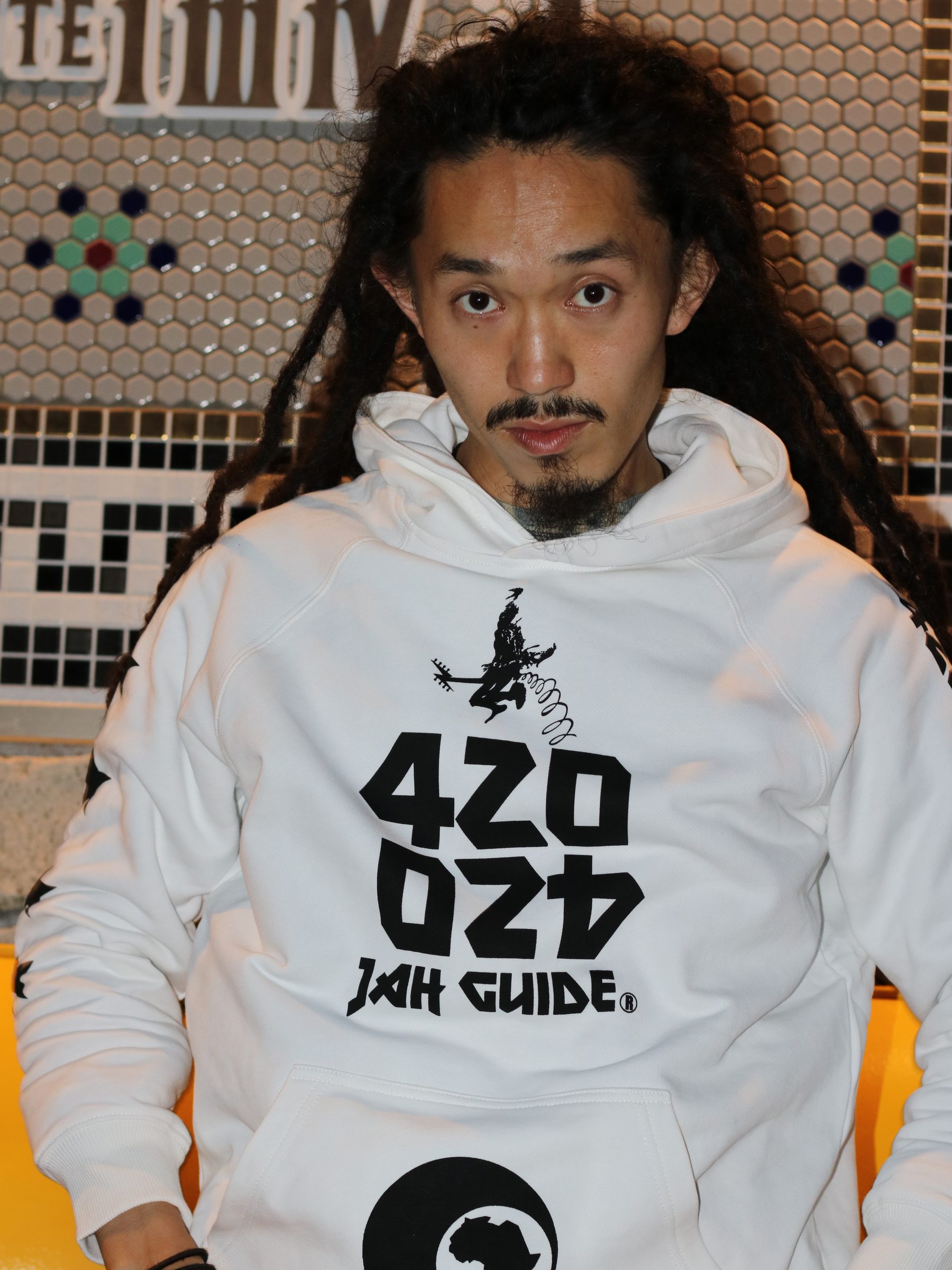 JAH GUIDEのロゴフーディを着た男性。タイル壁を背にした一枚